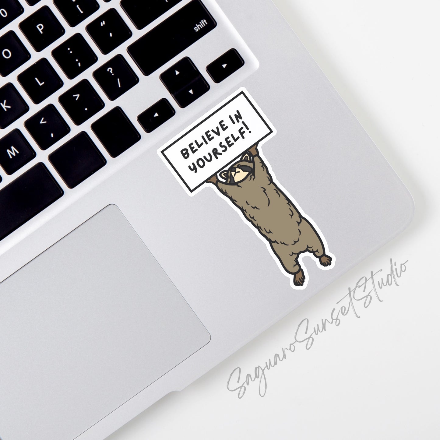 Sticker of a brown raccoon holding a white sign that says 'believe in yourself!'. The sticker is on the corner of a laptop.