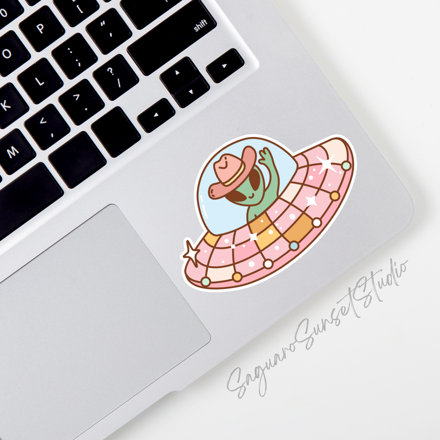 A sticker featuring a cute green alien wearing a cowboy hat, waving from inside a colorful and sparkly UFO. The UFO has a dome and is decorated with pastel colors and stars.