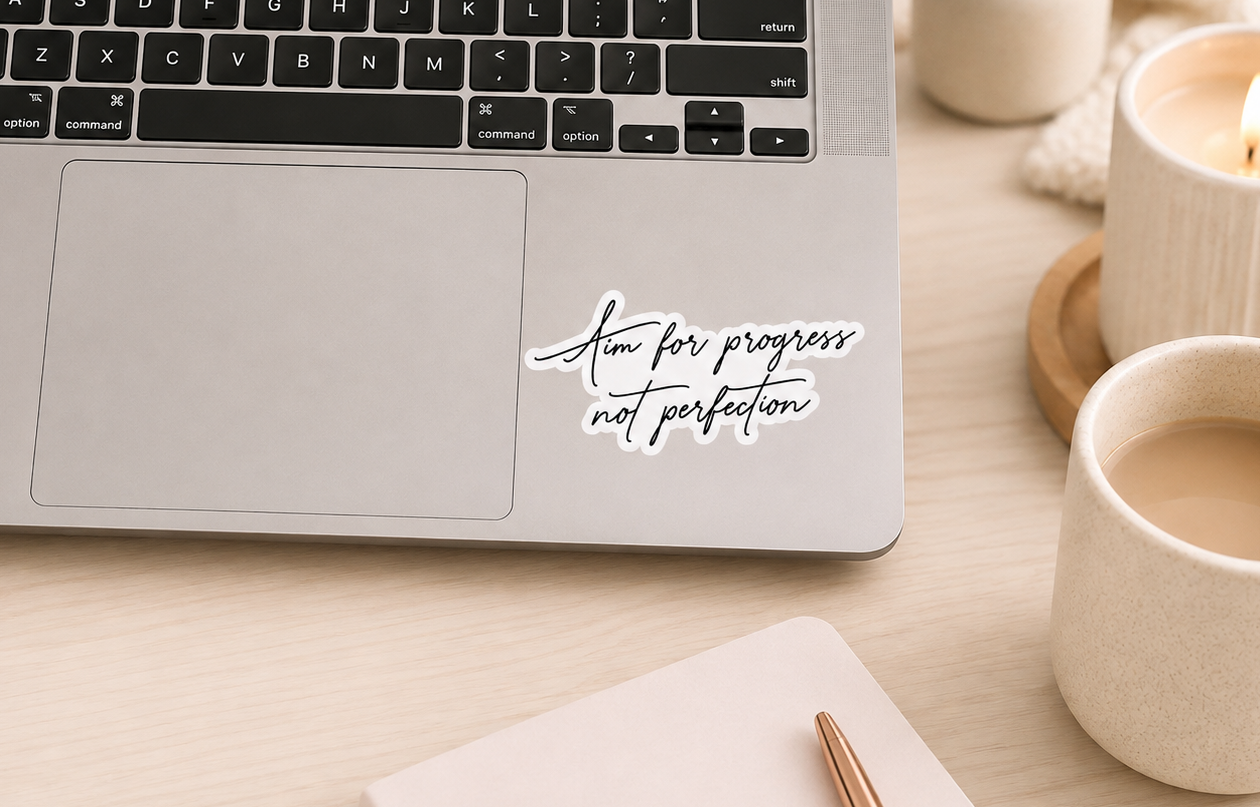A silver laptop on a light wood desk with a white script decal that reads ‘Aim for progress not perfection' on the trackpad. A notebook, pen, and two lit ceramic candles are arranged around the laptop in a warm, cozy workspace setting.