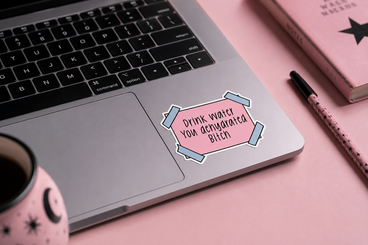 A pink die-cut sticker with the text 'Drink water you dehydrated Bitch' in black lettering, shown applied to the palm rest of a silver laptop on a pink desk with a mug, notebook, and pen nearby.
