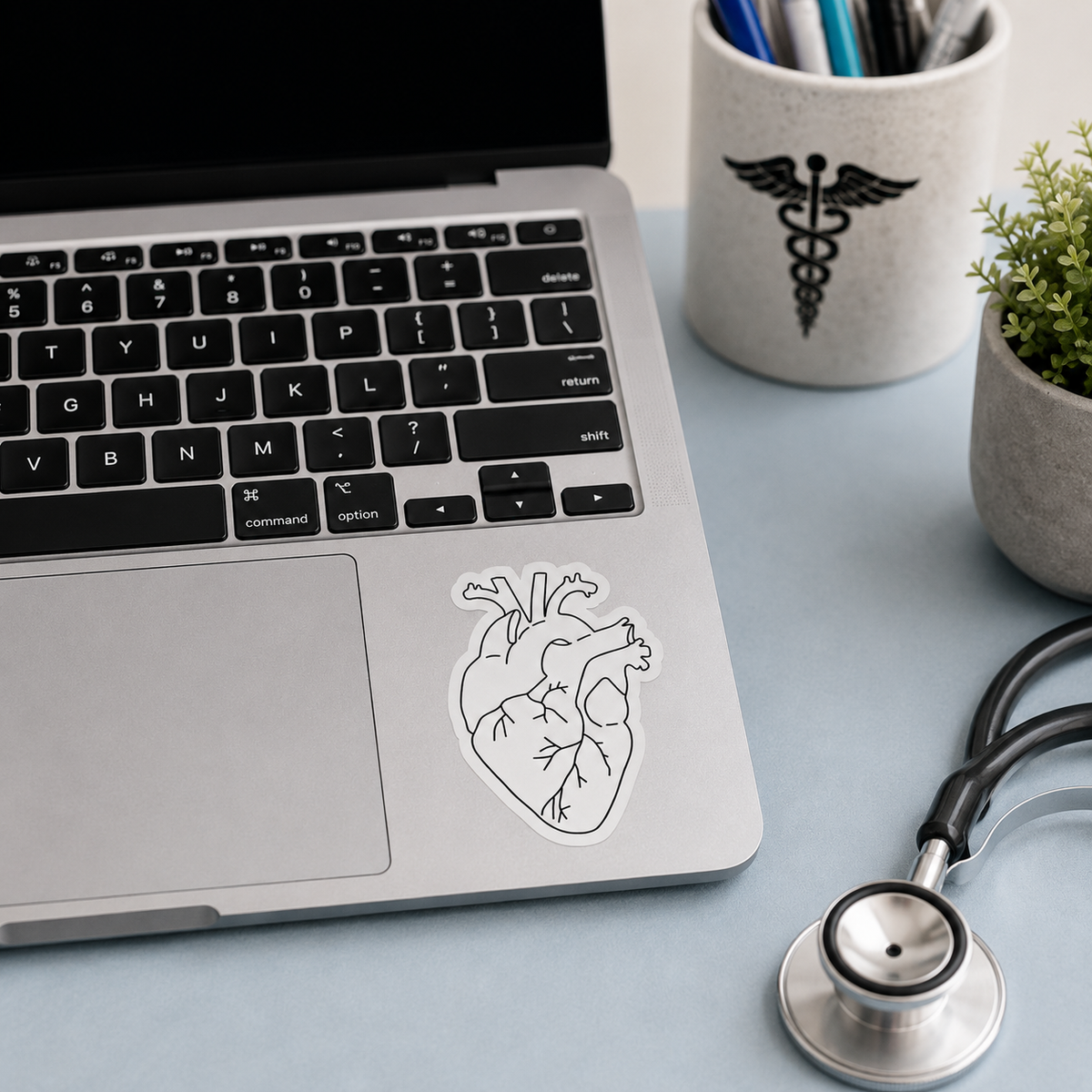 A die-cut vinyl sticker of a black and white anatomical heart on a silver laptop palm rest, styled on a light blue desk with a stethoscope, a laptop, a plant, and a medical cup.