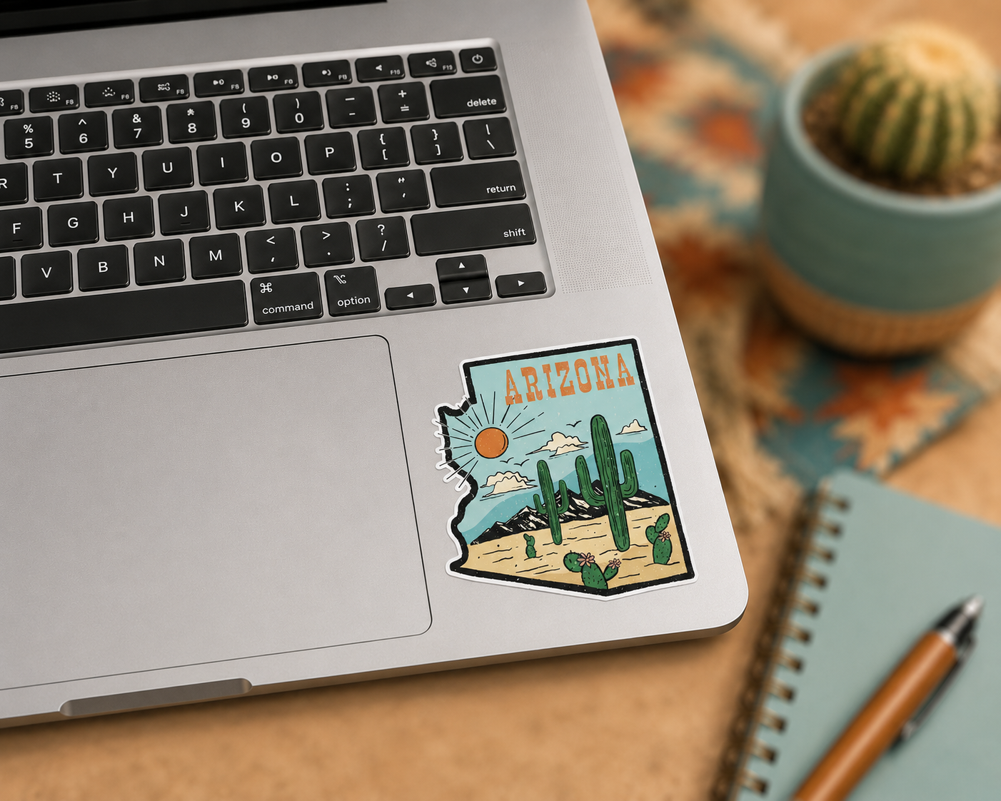A die-cut sticker shaped like the state of Arizona is applied to the lower right corner of a silver laptop. The sticker features a retro desert landscape with saguaro cacti, a sun, and the word 'ARIZONA' in orange.