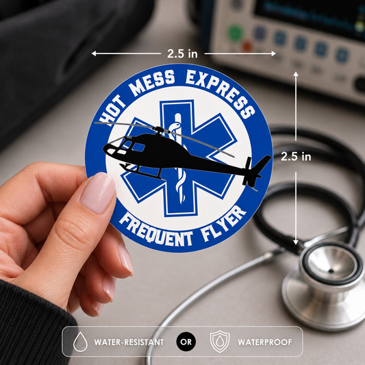 A hand holds a round blue and white sticker that reads 'HOT MESS EXPRESS' and 'FREQUENT FLYER' with a black helicopter over a medical-style star symbol. The sticker is shown with measurement arrows indicating 2.5 inches wide by 2.5 inches tall.