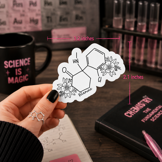 A hand holds a die-cut sticker featuring a chemical structure with floral details and the name ‘Ketamine'. The sticker is shown in a styled desk setting with dimension lines indicating 3.2 inches wide by 2.1 inches tall.