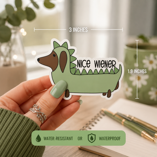 A hand holds a die-cut sticker shaped like a dachshund wearing a green dinosaur costume, with measurement lines showing 3 inches wide by 1.9 inches tall.