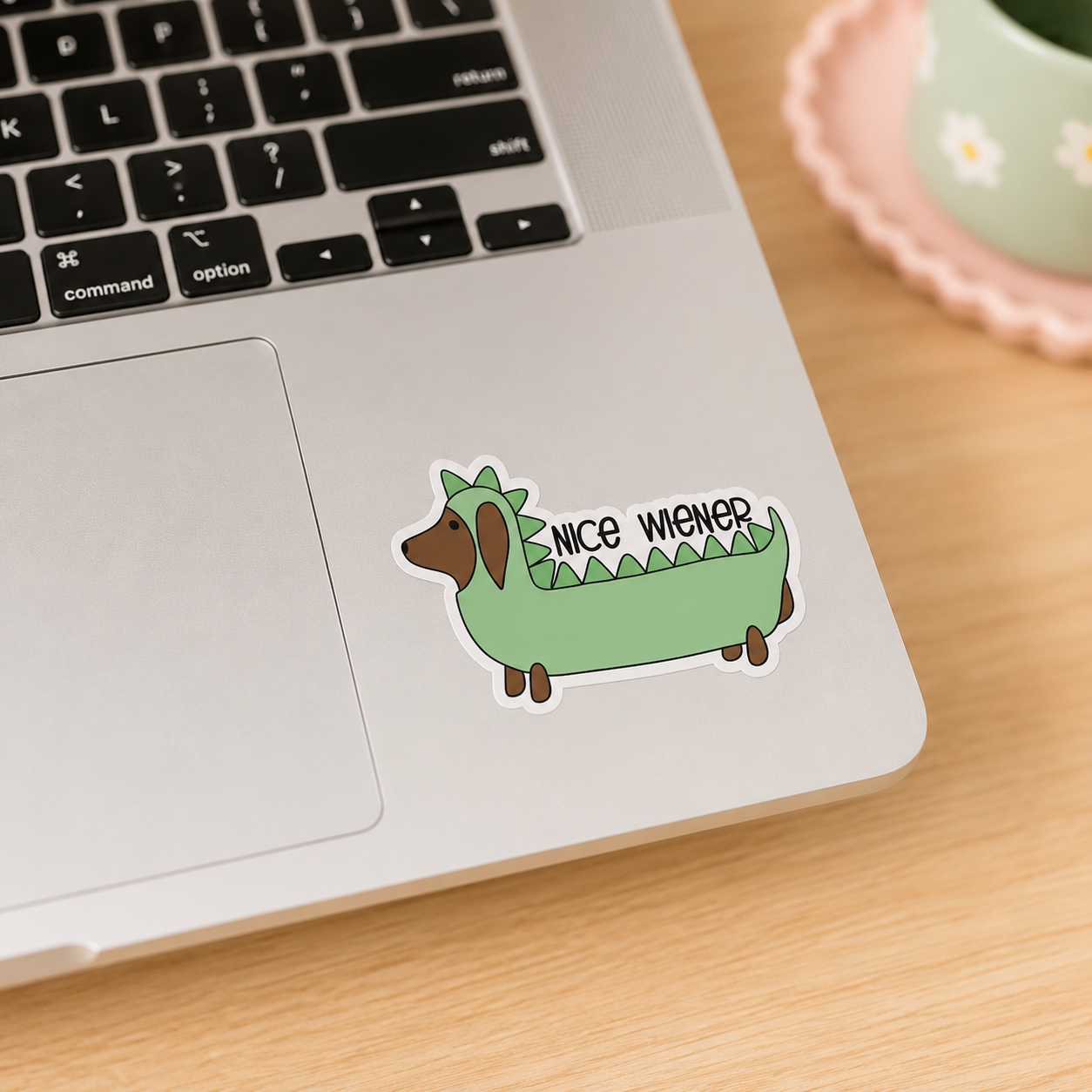 A die-cut sticker of a green dachshund wearing a dinosaur costume with the text 'NICE WIENER' on its back, shown applied to the palm rest of a silver laptop on a wooden desk next to a cup.