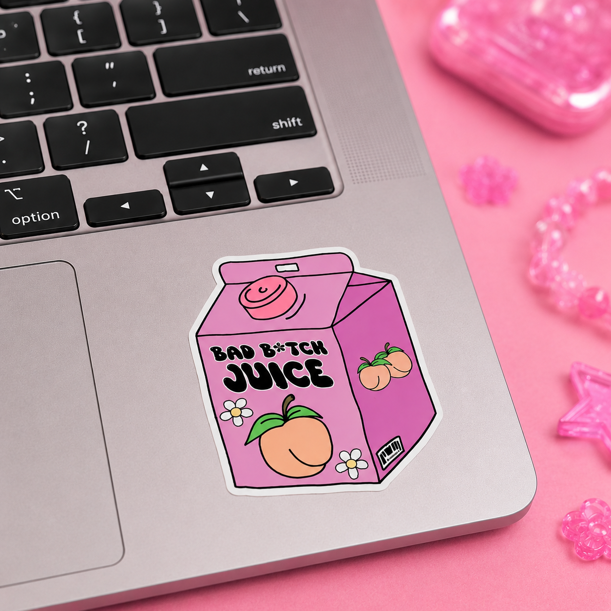 A die-cut sticker shaped like a pink juice carton with the text 'BAD B*TCH JUICE' and peach illustrations, placed on the palm rest of a silver laptop. The scene is styled on a pink surface with small pink accessories nearby.