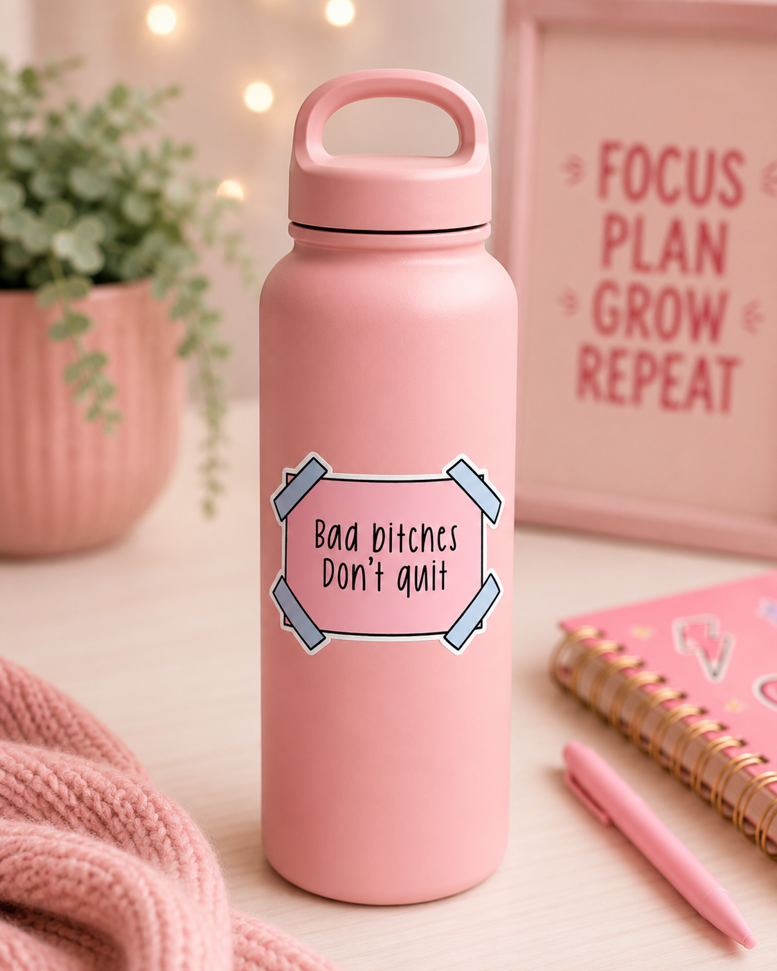 A pink insulated water bottle with a handle and a printed motivational quote sticker reading 'Bad bitches Don't quit' sits on a desk surrounded by a pink knit blanket, a notebook, a pen, and a potted plant.