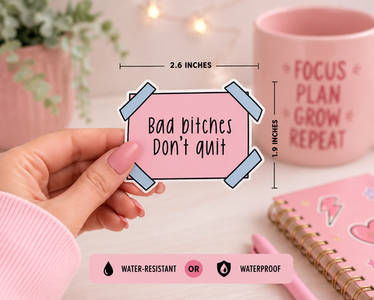 A hand holds a pink rectangular sticker with the text 'Bad bitches Don't quit' taped with blue tape graphics. The image includes dimension markers showing 2.6 inches wide by 1.9 inches tall