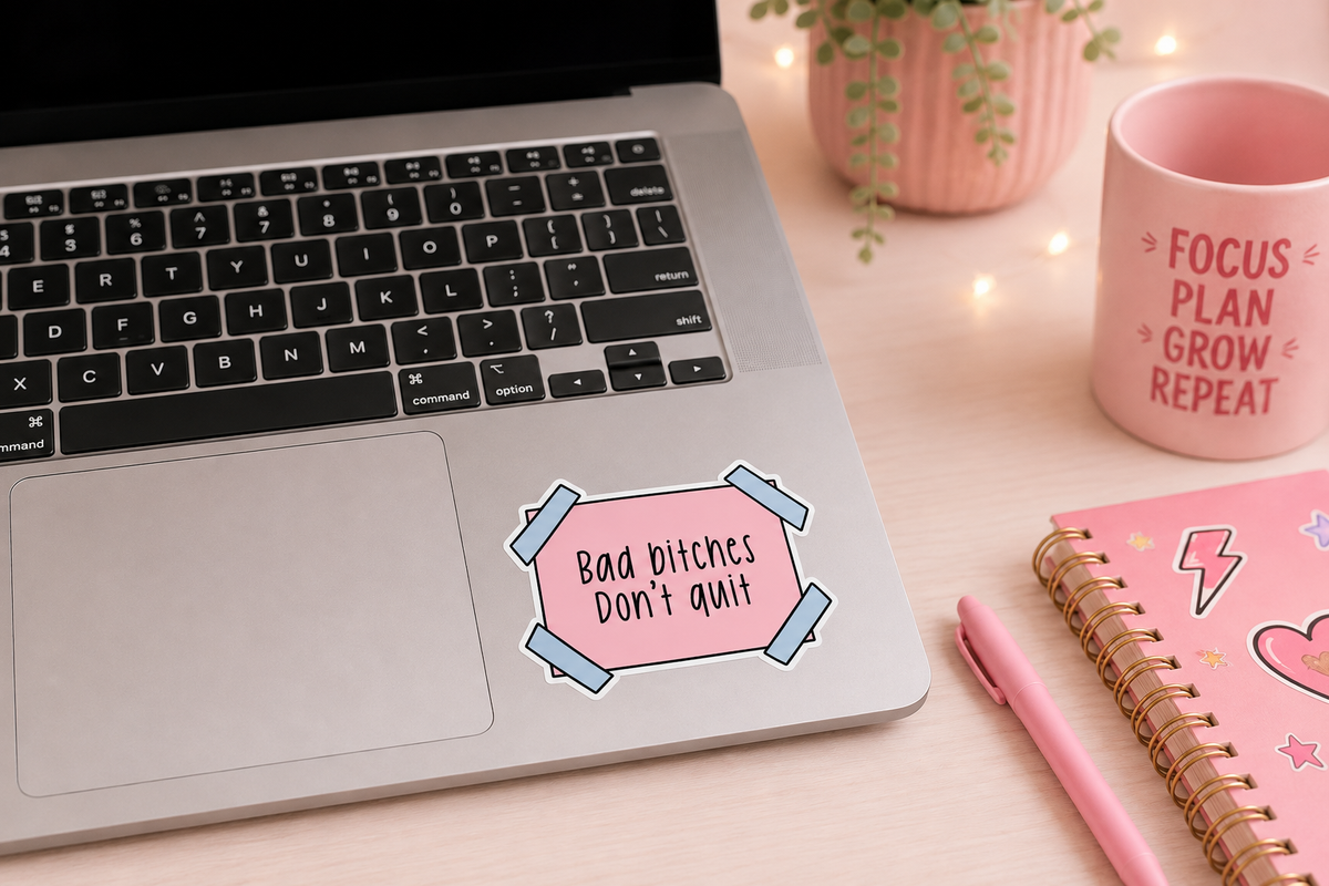 A pink rectangular sticker with the text 'Bad bitches Don't quit' in black script, styled on a silver laptop palm rest in a desk setting with a pink mug, notebook, and plant.