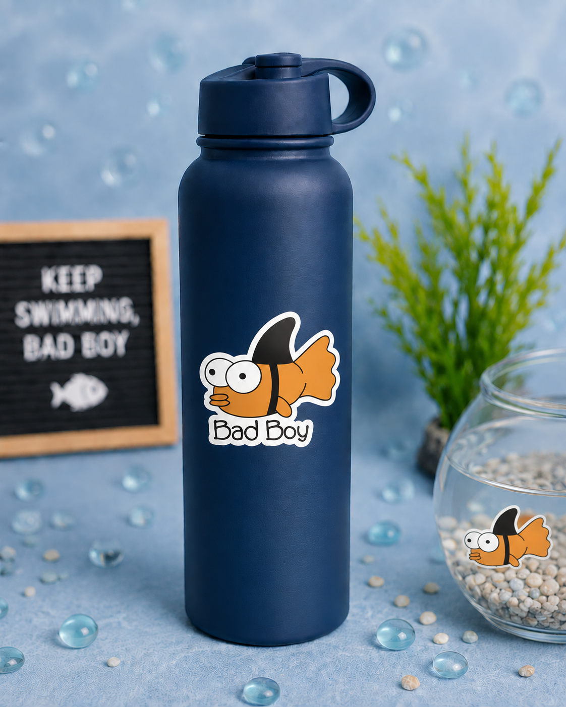 A navy blue insulated water bottle with a cartoon gold fish sticker that reads 'Bad Boy' is centered on a light blue surface. The scene includes a small sign reading 'KEEP SWIMMING, BAD BOY,' a green plant, and a glass bowl of pebbles with a matching fish sticker inside.
