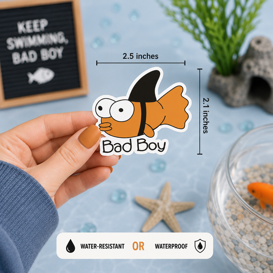 A hand holds a cartoon orange gold fish sticker with the words 'Bad Boy' and measurement lines showing 2.5 inches wide by 2.1 inches tall.