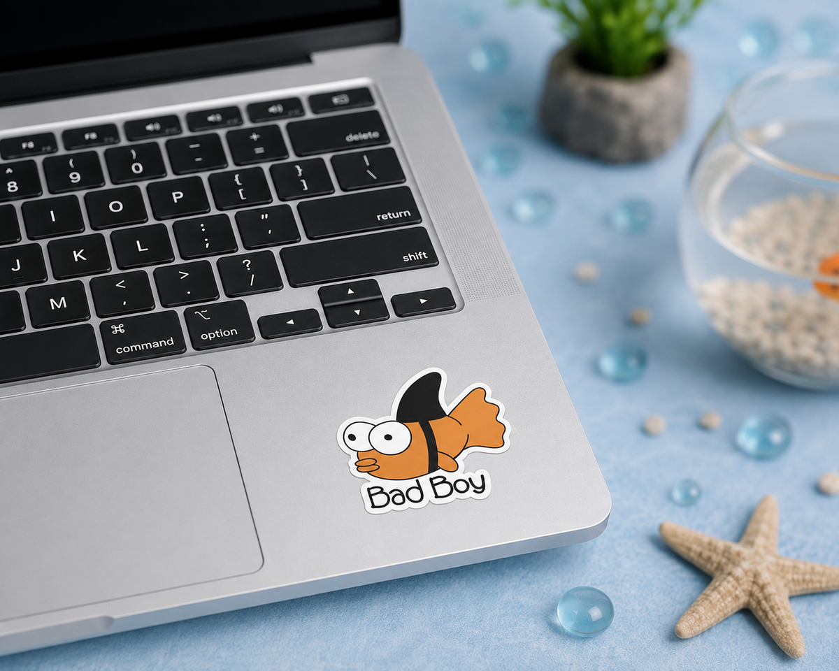 A cartoon gold fish sticker with the text 'Bad Boy' applied to the lower left corner of a silver laptop keyboard. The scene is styled as a flat-lay on a light blue surface with decorative props including a small potted plant, a glass bowl, and a starfish.