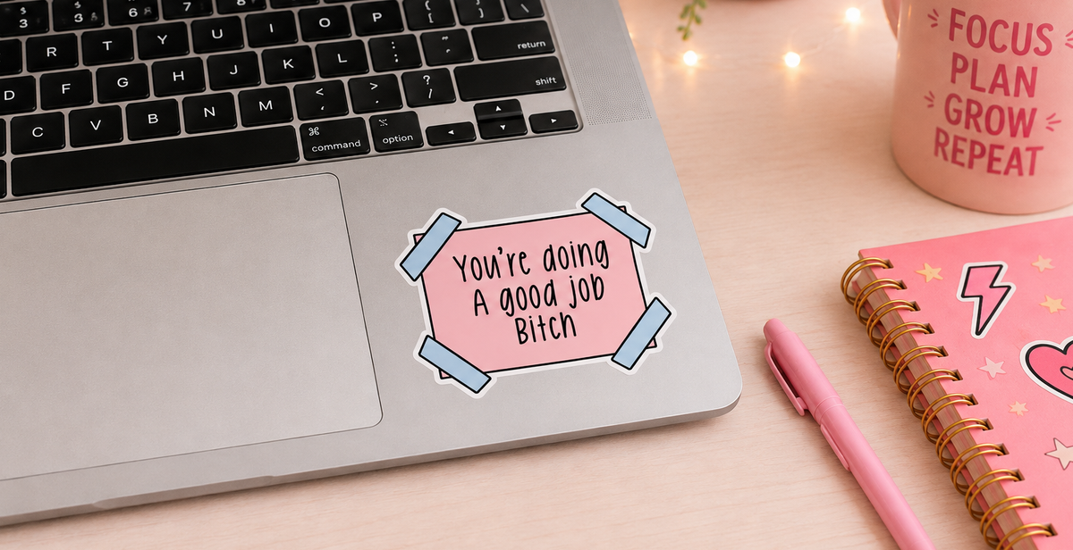 A pink sticker with the text 'You're doing A good Job Bitch' in black lettering, decorated with light blue tape graphics, shown applied to the palm rest of a silver laptop on a wooden desk. The scene is styled with a pink mug, a pink notebook, and a pink pen.