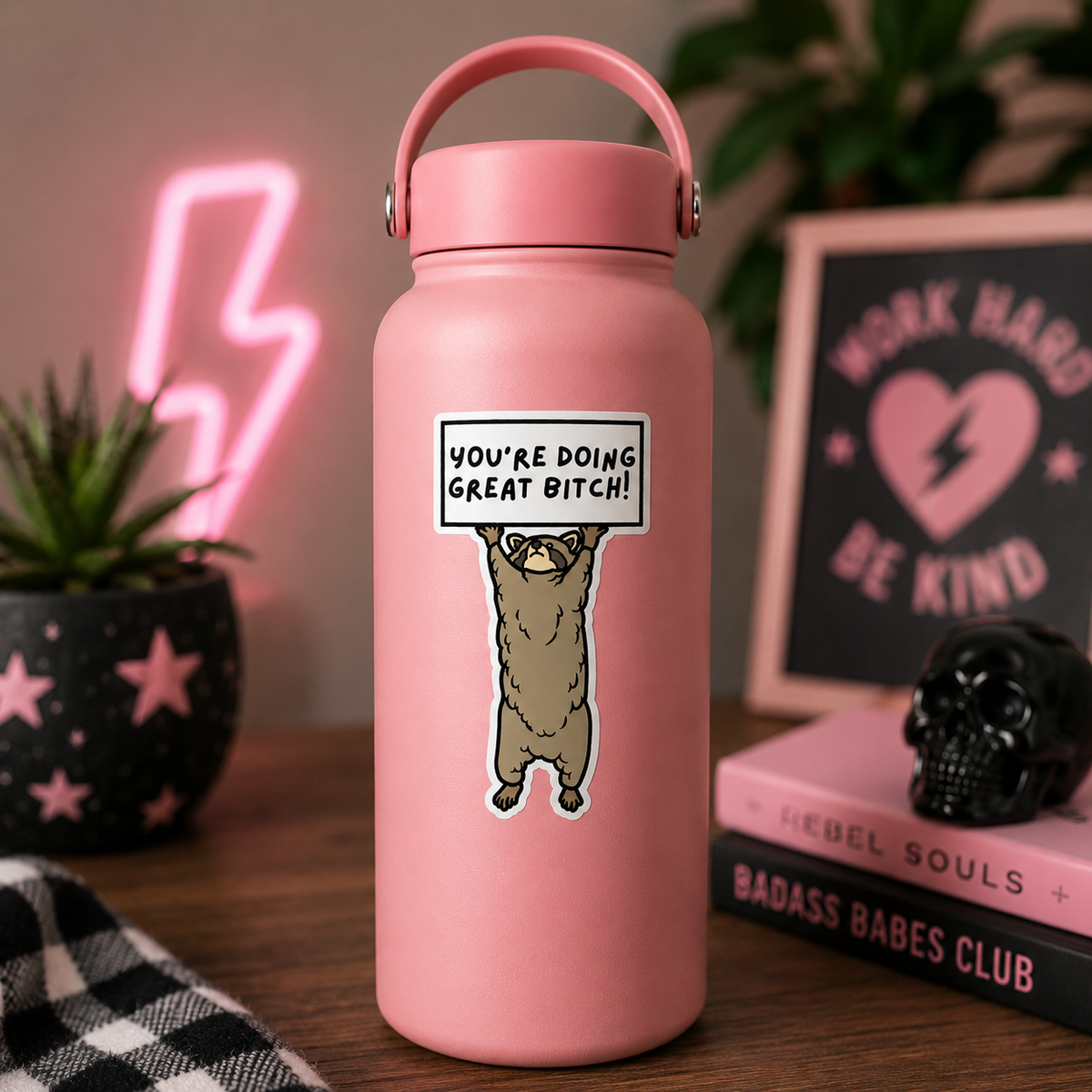 A pink insulated water bottle with a handle and a cartoon raccoon holding a sign that reads 'YOU'RE DOING GREAT BITCH!' in a styled indoor setting with plants, books, and a neon light in the background.