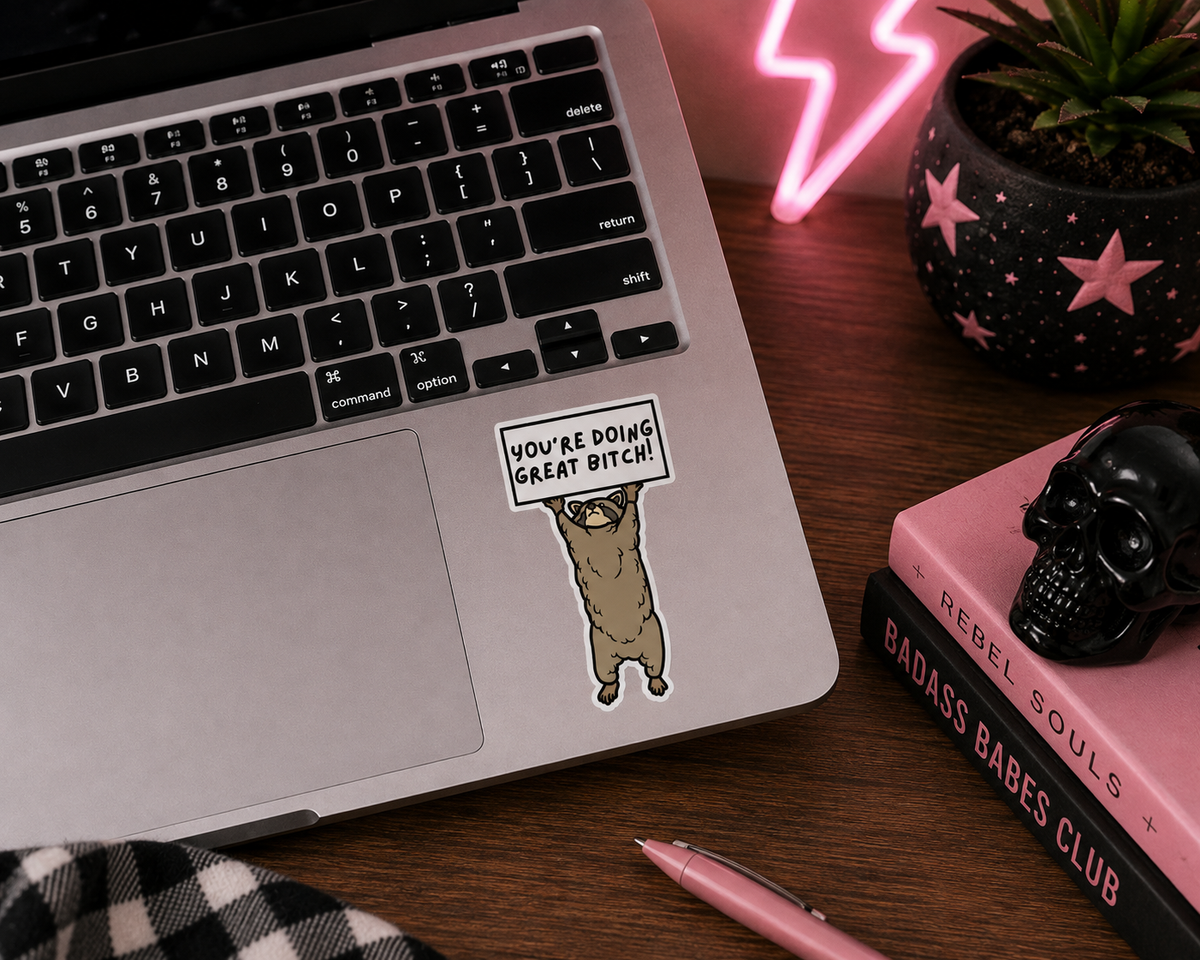 A die-cut sticker featuring a cartoon raccoon holding a sign that reads 'YOU'RE DOING GREAT BITCH!' applied to the palm rest of a silver laptop. The laptop is part of a styled desk flat-lay with a pink pen, a pink book, a black skull, and a glowing pink neon lamp.