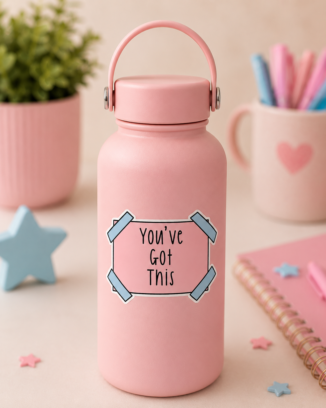 A pink insulated water bottle with a carrying loop lid, featuring a sticker that reads 'You've Got This' in black text inside a pink and blue outlined box. The bottle is set on a desk with a blurred plant, pens, and notebooks in the background.