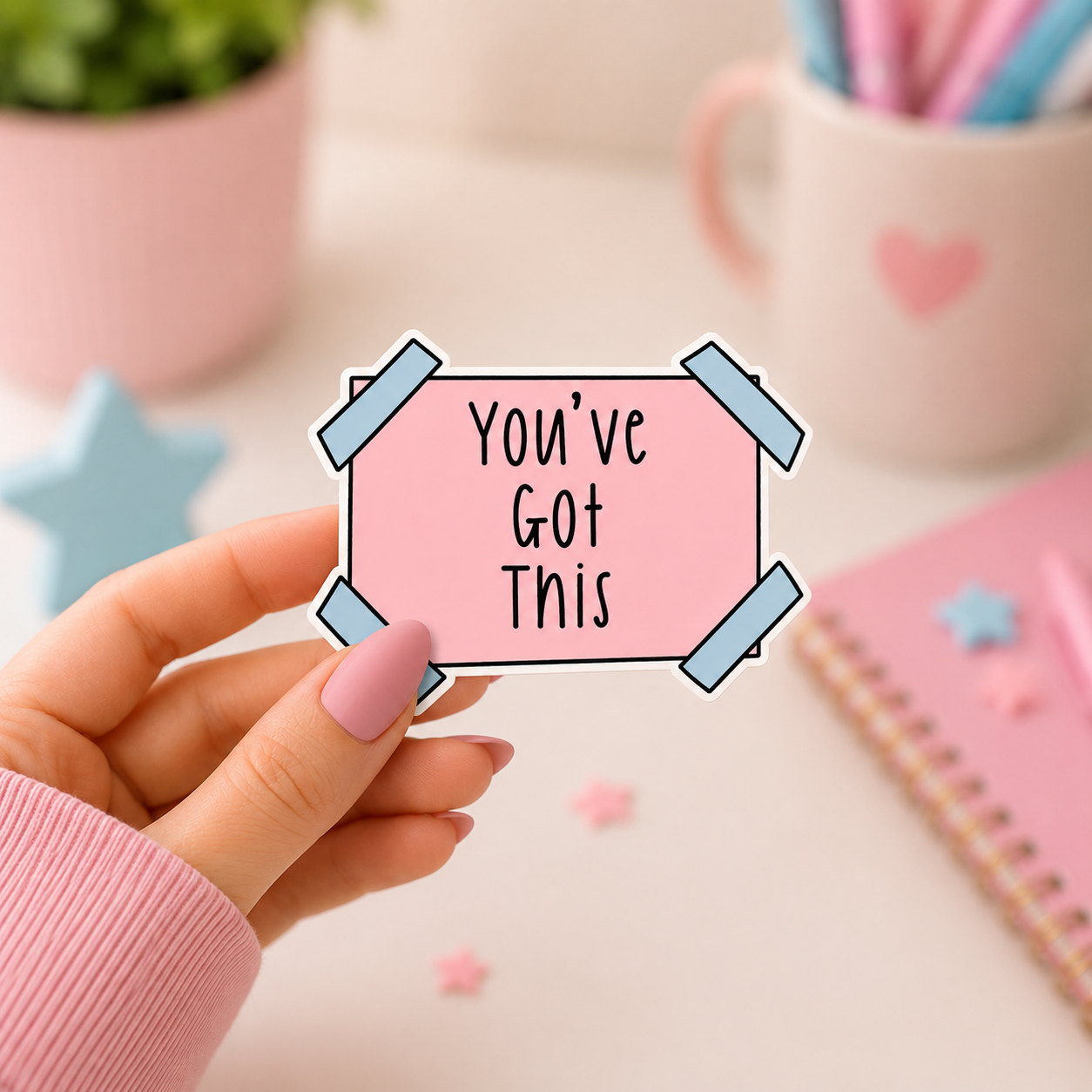 A hand with pink nails holds a pink sticker that reads 'You've Got This' with light blue tape graphics, set against a cute desk background with a pink notebook, mug, and plant.