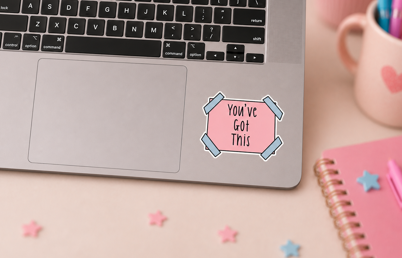 A pink die-cut sticker with the text 'You've Got This' in black, styled on the palm rest of a silver laptop. The scene is a styled desk flat-lay with a pink notebook, a pink mug, and pastel star decorations.