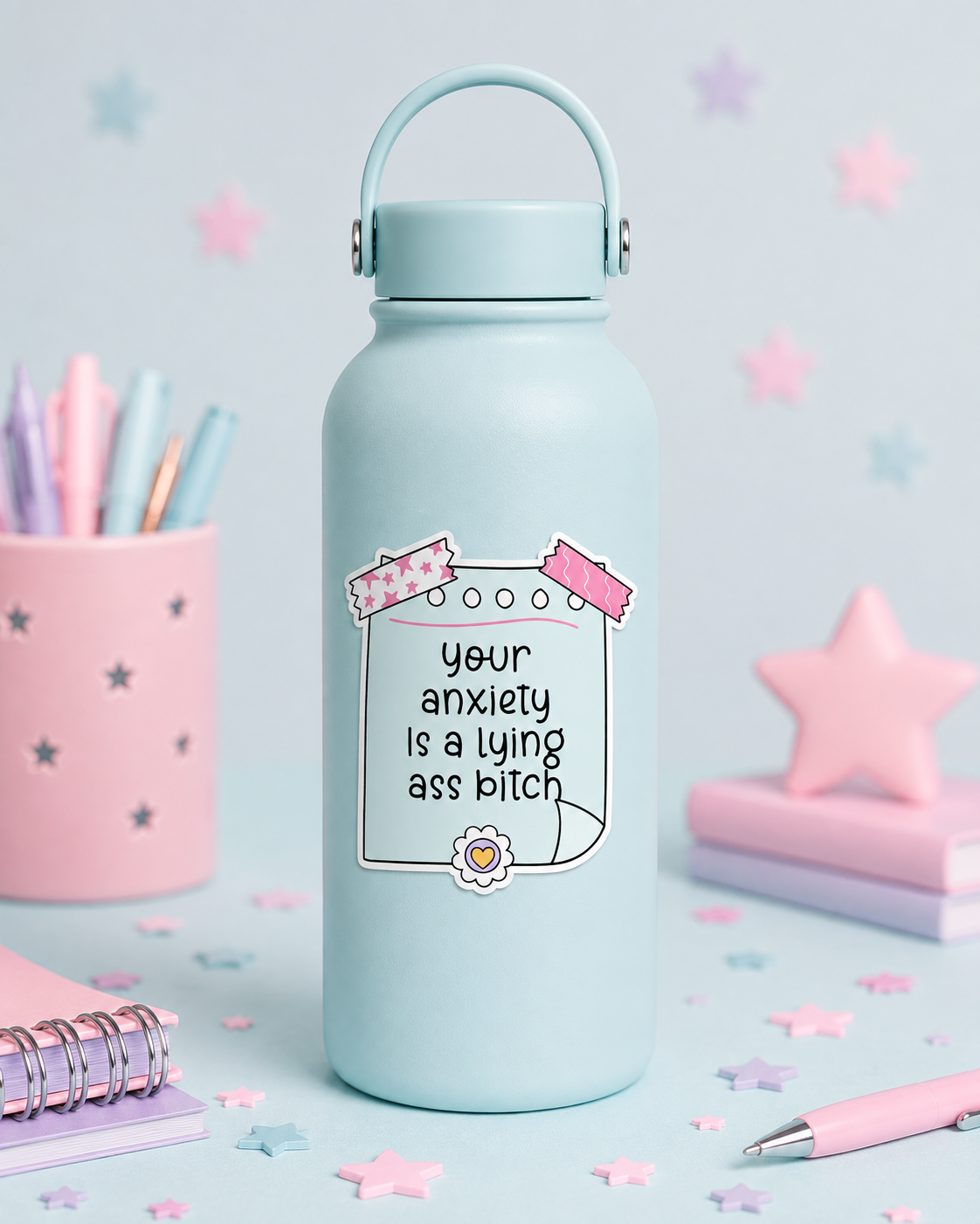 A light blue insulated water bottle with a carrying loop lid, featuring a printed graphic that reads 'your anxiety is a lying ass bitch' with pink ribbon and flower doodles. The bottle is styled on a pastel desk with pink star confetti, a pink pen cup, notebooks, and a pink star-shaped object in the background.