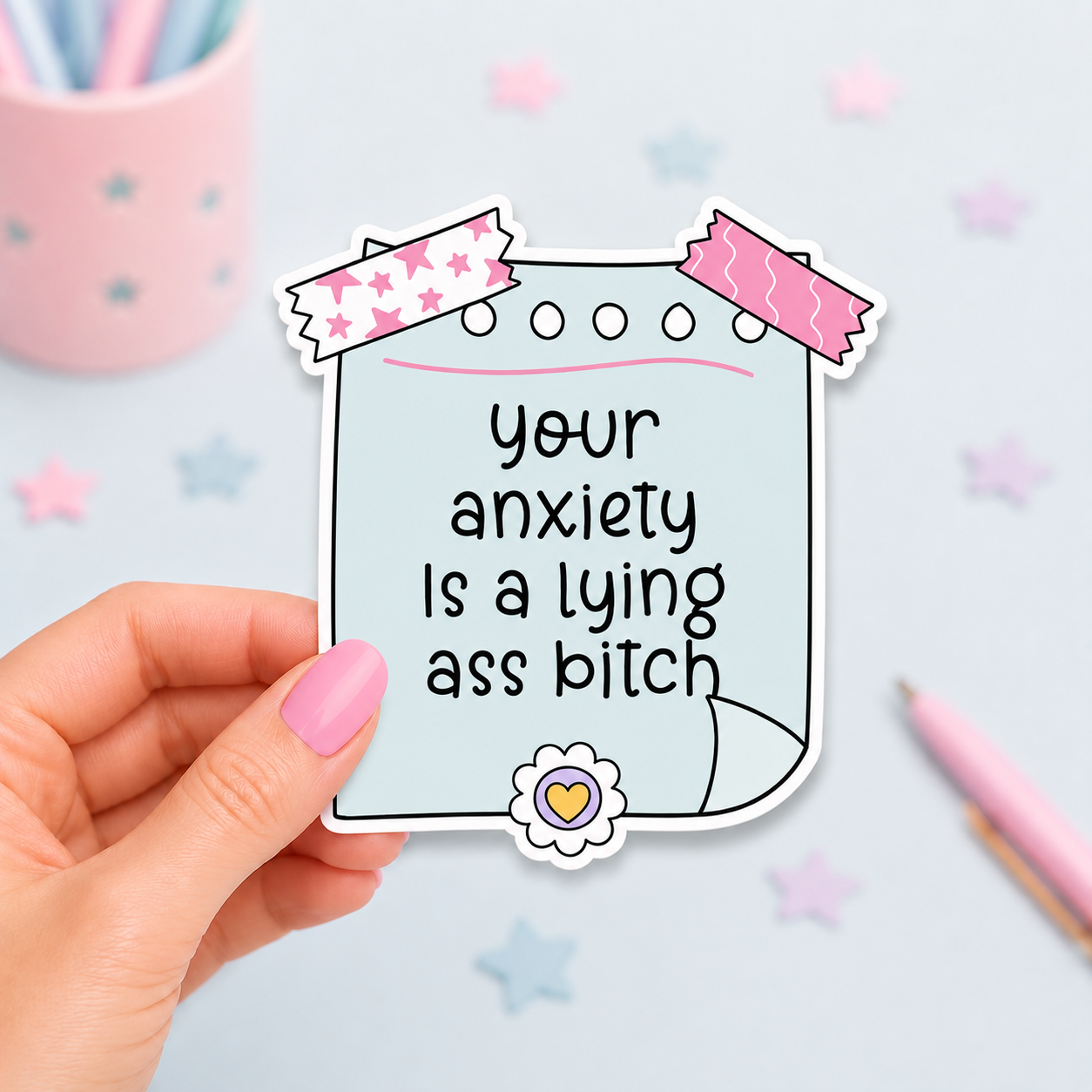 A hand with pink nail polish holds a die-cut sticker shaped like a piece of paper with the text 'your anxiety Is a lying ass bitch' in black lettering. The sticker has a light blue background, decorated with pink and white patterned tape at the top and a small flower with a yellow heart at the bottom.