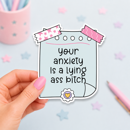 A hand with pink nail polish holds a die-cut sticker shaped like a piece of paper with the text 'your anxiety Is a lying ass bitch' in black lettering. The sticker has a light blue background, decorated with pink and white patterned tape at the top and a small flower with a yellow heart at the bottom.