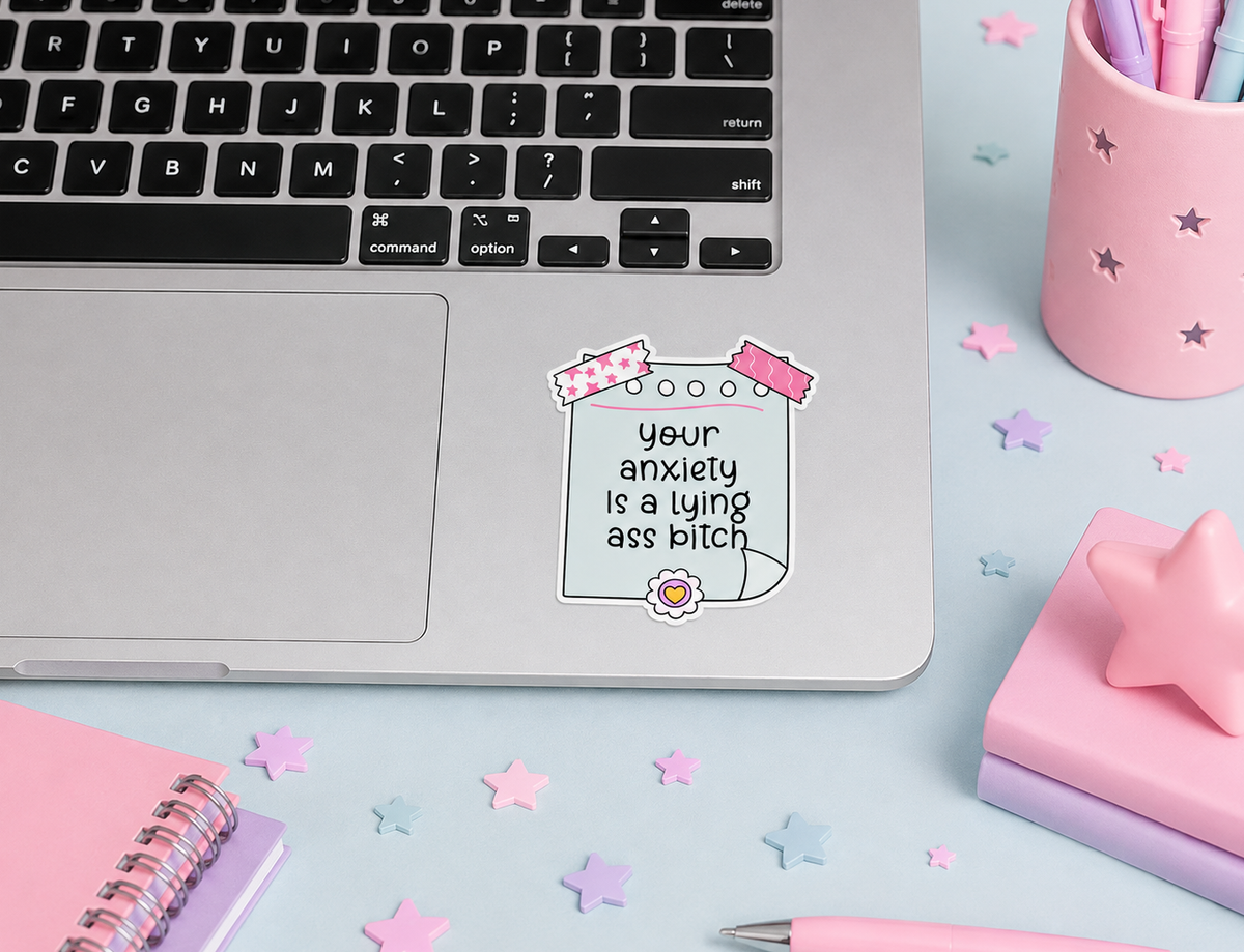 A square die-cut sticker with the text 'Your anxiety is a lying ass bitch' in black lettering, decorated with pink patterned ribbon graphics and a small flower, shown applied to the palm rest of a silver laptop on a pastel desk setup.