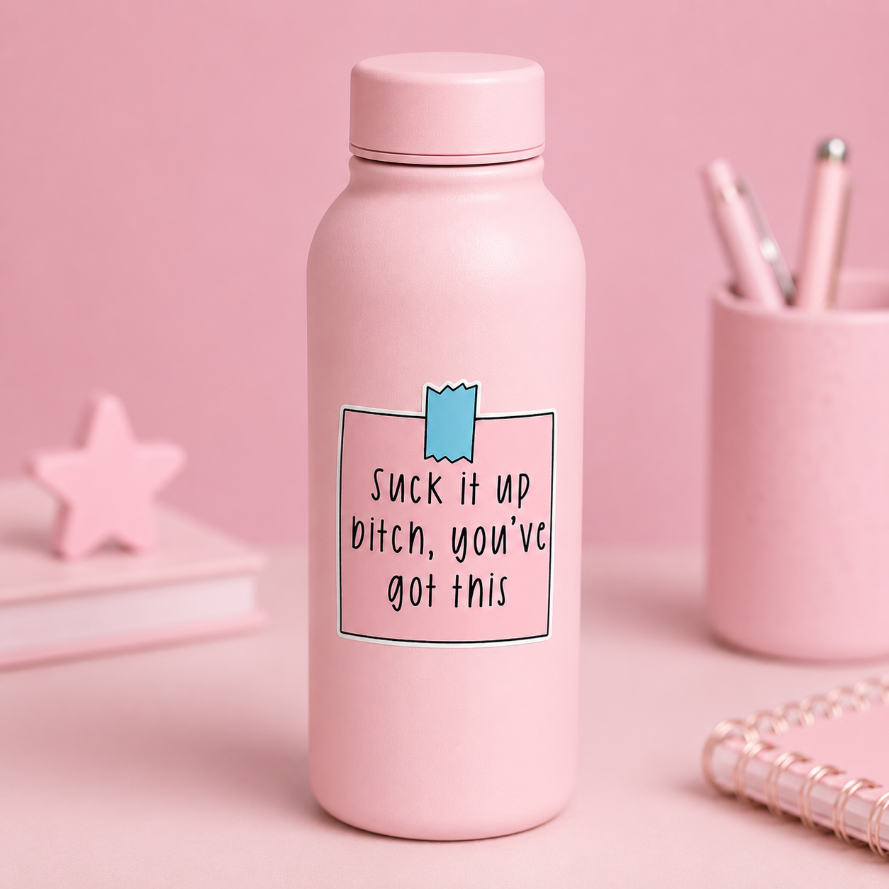 A light pink insulated water bottle with a matching lid, featuring a printed graphic on the front with the text 'Suck it up bitch, you've got this' in black lettering inside a thin black box, with a small blue ribbon detail above. The bottle is set on a pink surface with a pink background, styled with a pink star decoration, a pencil cup with pens, and a notebook.