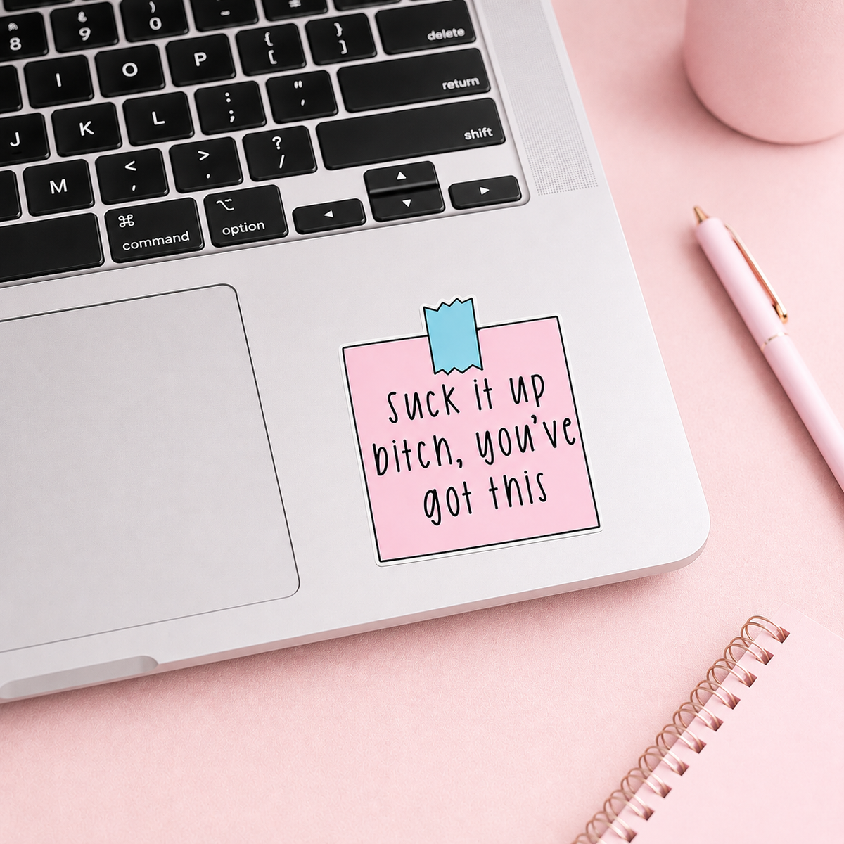 A pink sticky note decal with the text 'Suck it up bitch, you've got this' in black handwritten-style lettering, attached to a silver laptop on a pink desk with a pen, notebook, and mug visible.