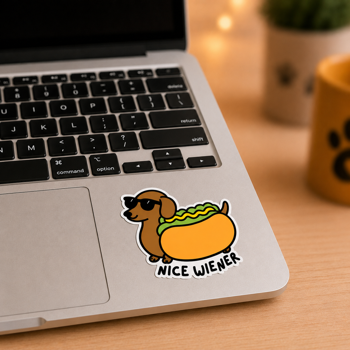 A die-cut vinyl sticker of a dachshund wearing sunglasses with a hotdog and the text 'NICE WIENER' applied to the bottom left corner of a silver laptop. The laptop is shown on a wooden desk with a warm, blurred background containing a plant and a yellow mug.