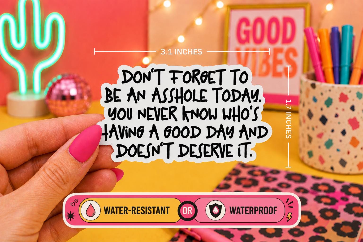 A hand with pink nail polish holds a die-cut sticker with a humorous quote ‘Don’t forget to be an asshole today. You never know who’s having a good day and doesn’t deserve it'. The sticker is shown in a colorful, styled desk setting with dimension lines indicating it is 3.1 inches wide by 1.7 inches tall.