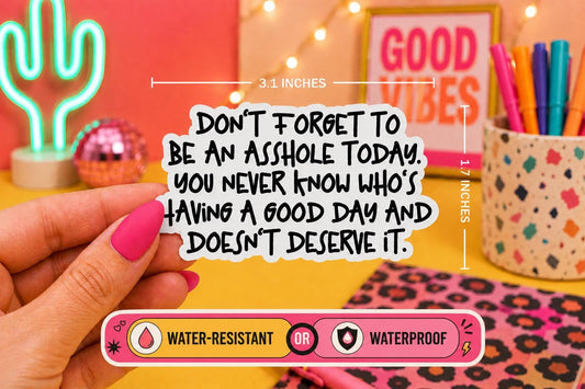 A hand with pink nail polish holds a die-cut sticker with a humorous quote ‘Don’t forget to be an asshole today. You never know who’s having a good day and doesn’t deserve it'. The sticker is shown in a colorful, styled desk setting with dimension lines indicating it is 3.1 inches wide by 1.7 inches tall.
