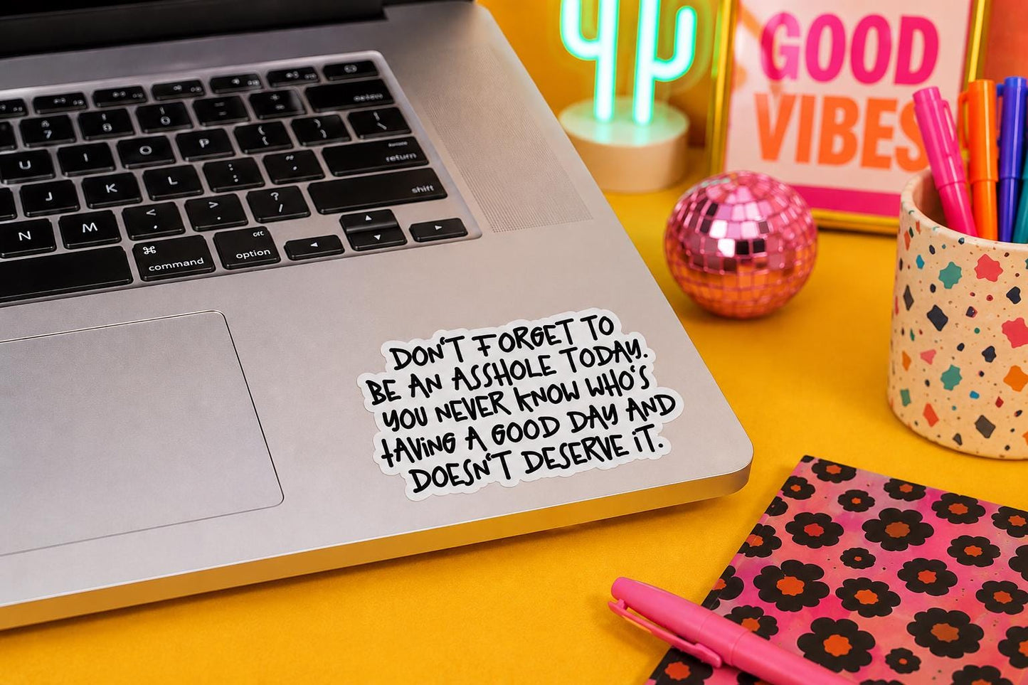 A silver laptop on a bright yellow desk with a hand-written sticker that reads, 'DON'T FORGET TO BE AN ASSHOLE TODAY. YOU NEVER KNOW WHO'S HAVING A GOOD DAY AND DOESN'T DESERVE IT.'
