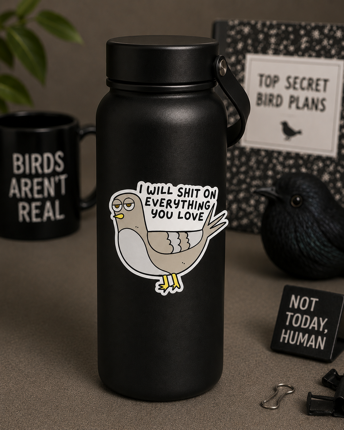 A matte black insulated water bottle with a cartoon pigeon graphic and the text 'I WILL SHIT ON EVERYTHING YOU LOVE' on the front, styled on a tabletop with a black mug, bird figurine, and other desk props.