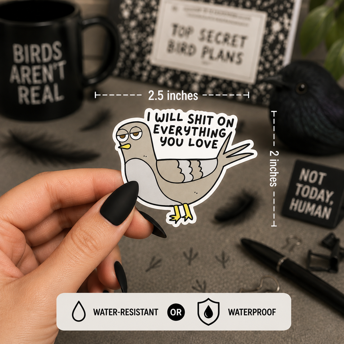 A hand with black nail polish holds a die-cut sticker of a grumpy pigeon holding a sign that reads 'I WILL SHIT ON EVERYTHING YOU LOVE'. The background is a styled desk with bird-themed items, including a black mug that says 'BIRDS AREN'T REAL', a bird figurine, and a notebook.