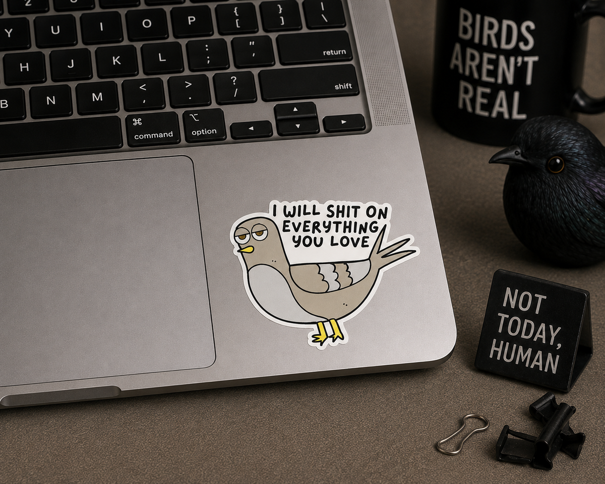 A die-cut sticker featuring a cartoon pigeon with glasses and the text 'I WILL SHIT ON EVERYTHING YOU LOVE' applied to the palm rest of a silver laptop. The scene is styled as a desk flat-lay with a black mug that reads 'BIRDS AREN'T REAL', a black bird figurine, and a small black sign that says 'NOT TODAY, HUMAN'.