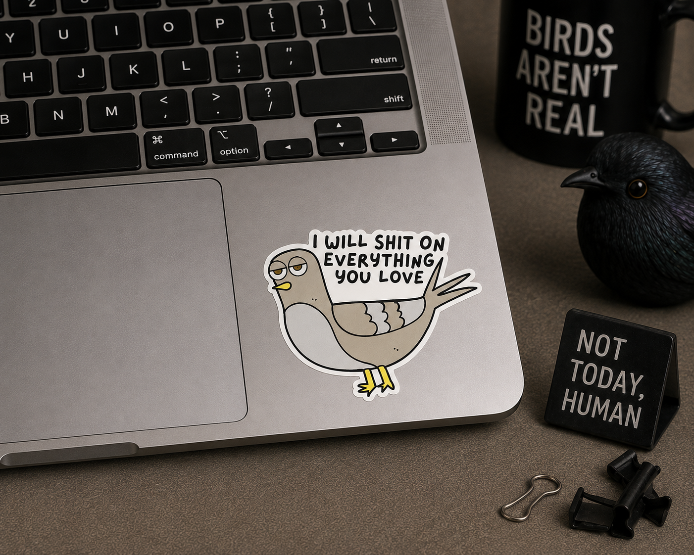 A die-cut sticker featuring a cartoon pigeon with glasses and the text 'I WILL SHIT ON EVERYTHING YOU LOVE' applied to the palm rest of a silver laptop. The scene is styled as a desk flat-lay with a black mug that reads 'BIRDS AREN'T REAL', a black bird figurine, and a small black sign that says 'NOT TODAY, HUMAN'.