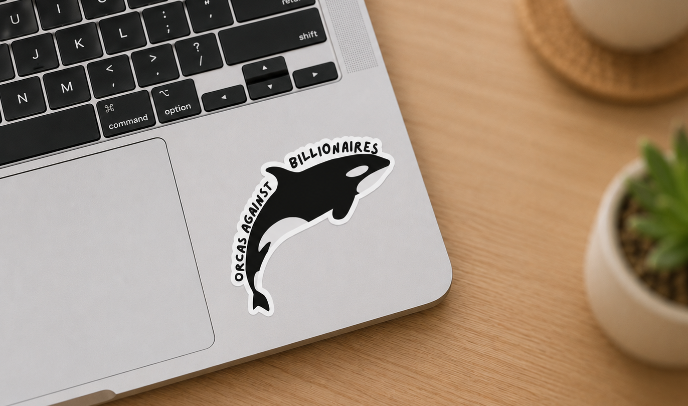 A die-cut vinyl sticker of a black and white orca with the text 'ORCAS AGAINST BILLIONAIRES' applied to the palm rest of a silver laptop on a wooden desk.