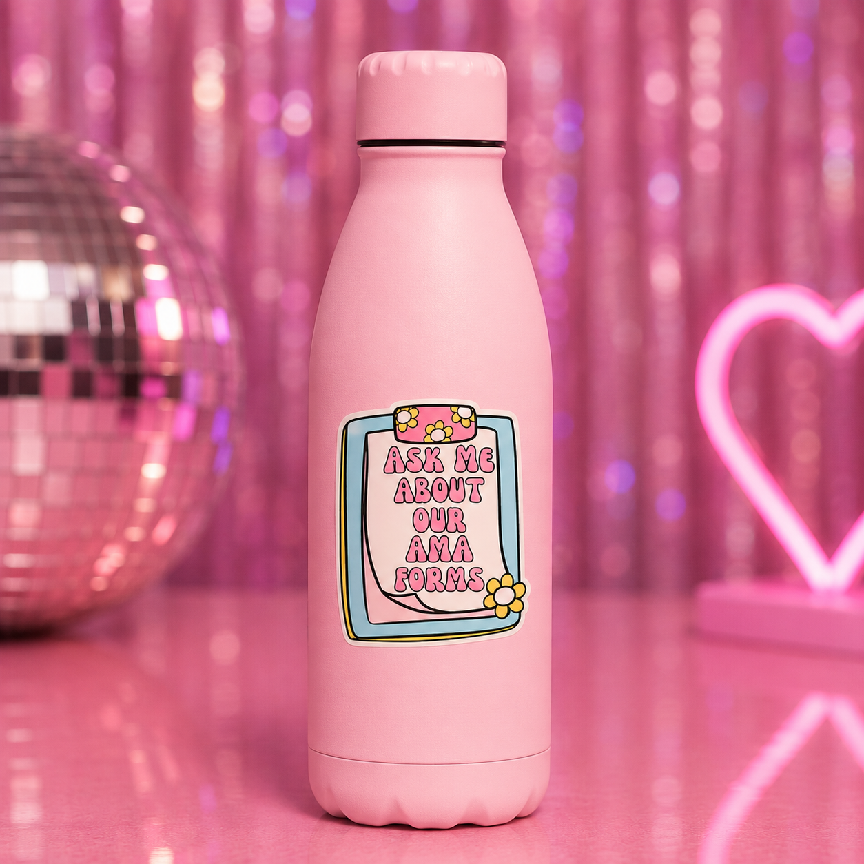 A pink insulated bottle with a matching pink cap, featuring a cartoon-style label that reads 'ASK ME ABOUT OUR AMA FORMS' with flowers and a blue border. The bottle is set on a pink reflective surface with a disco ball on the left and a pink neon heart on the right against a pink tinsel backdrop.