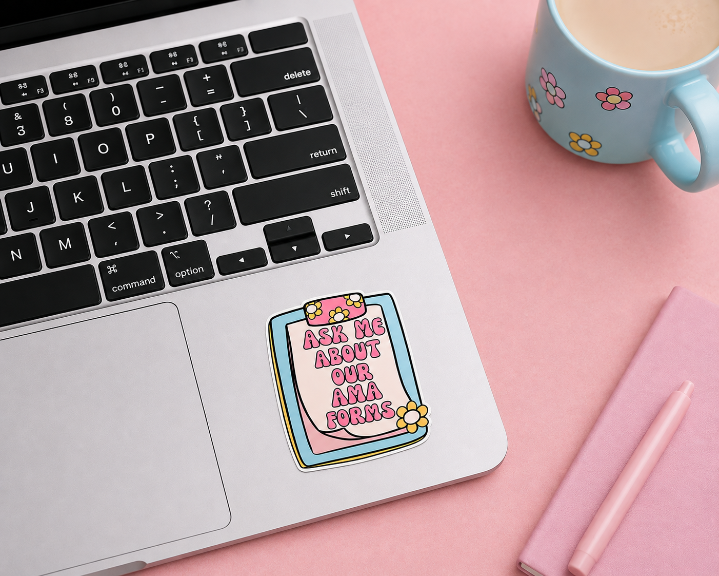 A die-cut sticker with the text 'ASK ME ABOUT OUR AMA forms in pink lettering on a pink clipboard graphic, shown applied to the palm rest of a silver laptop. The scene is styled on a pink surface with a light blue floral mug and a pink notebook in the background.