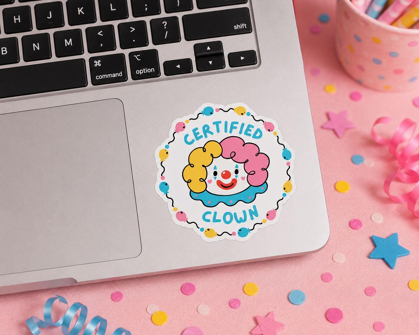 A round die-cut sticker featuring a cartoon clown with yellow and pink hair and the words 'CERTIFIED CLOWN' in blue, shown applied to the lower right corner of a silver laptop on a pink confetti-covered surface.