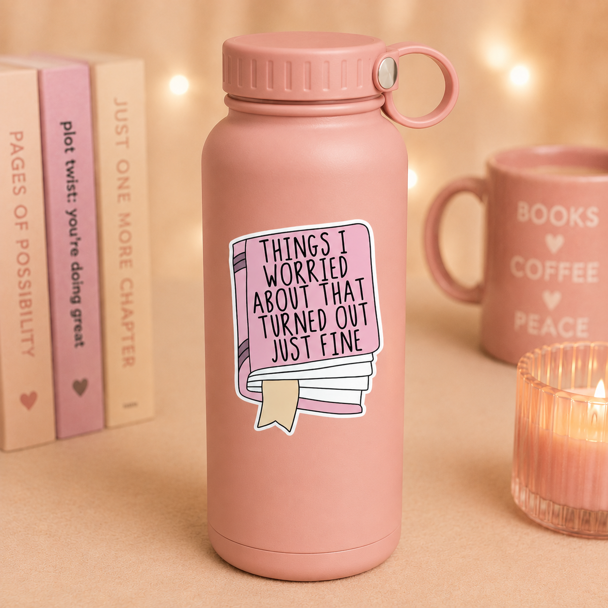 A dusty pink insulated water bottle with a matching lid and carry loop, featuring a sticker that reads 'THINGS I WORRIED ABOUT THAT TURNED OUT JUST FINE' with a book illustration. The bottle is styled on a beige surface with warm bokeh lights, a pink mug reading 'BOOKS COFFEE PEACE', and books in the background.