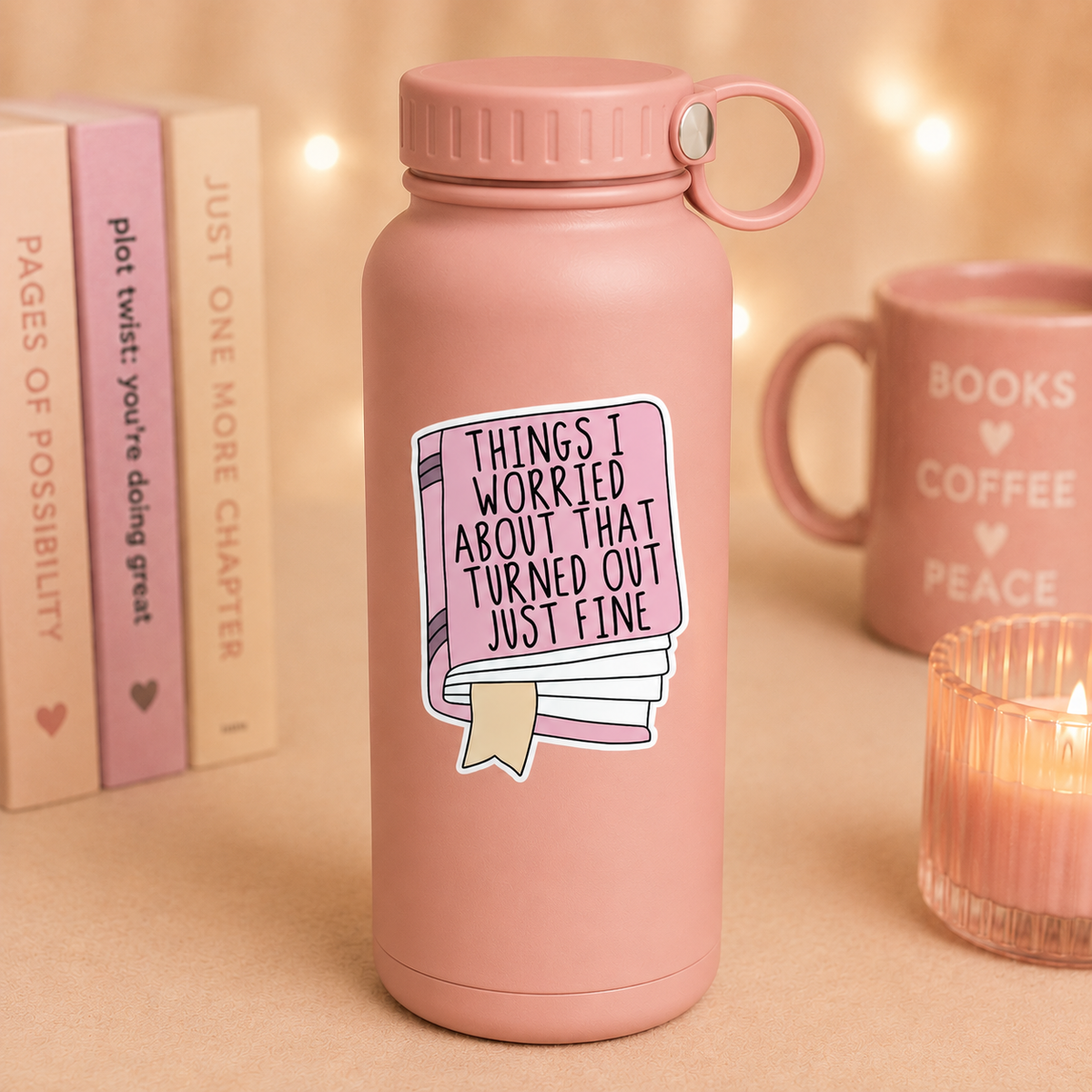 A dusty pink insulated water bottle with a matching lid and carry loop, featuring a sticker that reads 'THINGS I WORRIED ABOUT THAT TURNED OUT JUST FINE' with a book illustration. The bottle is styled on a beige surface with warm bokeh lights, a pink mug reading 'BOOKS COFFEE PEACE', and books in the background.
