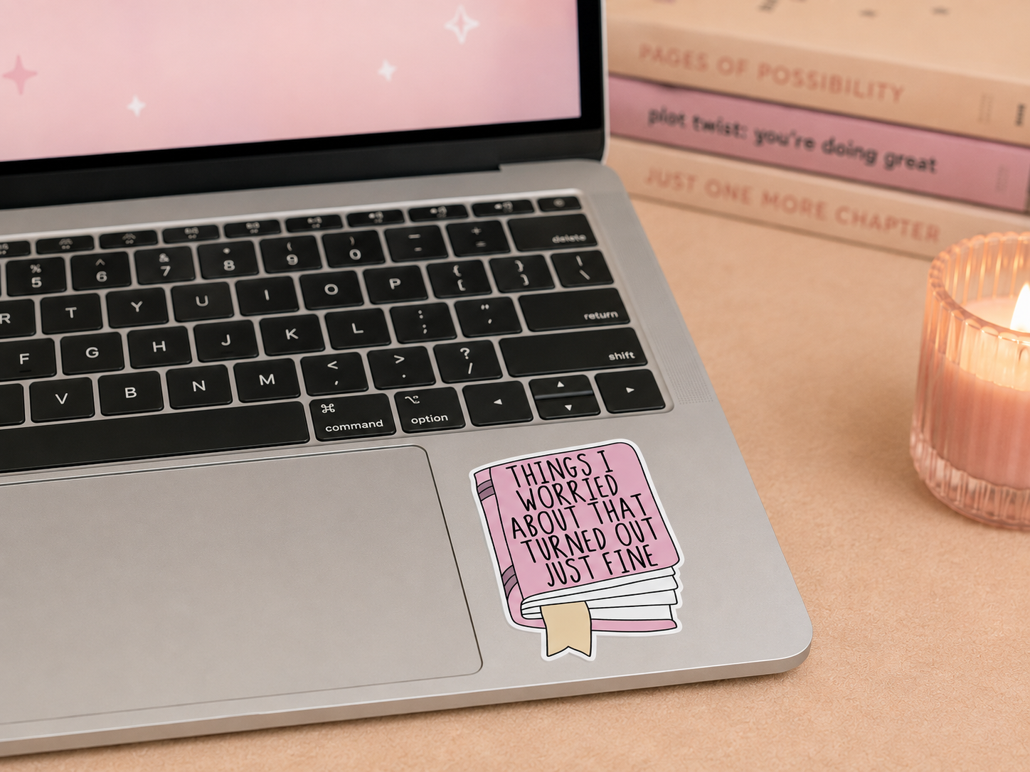 A pink book-shaped sticker with the text 'THINGS I WORRIED ABOUT THAT TURNED OUT JUST FINE' applied to the palm rest of a silver laptop. The laptop sits on a beige desk next to a lit pink candle and stacked books in the background.
