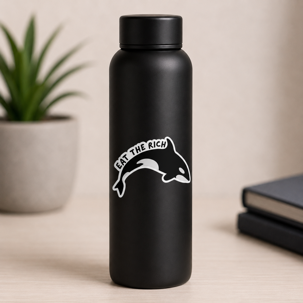 A matte black insulated water bottle with a white graphic of an orca and the text 'EAT THE RICH' on the front, standing on a light wood surface with a blurred potted plant and books in the background.