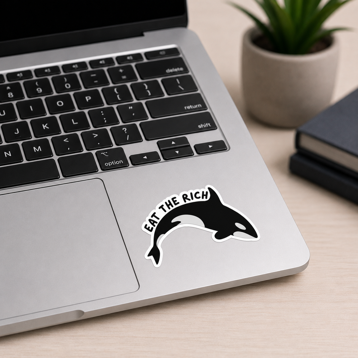 A black and white sticker featuring an orca and the text 'EAT THE RICH' applied to the palm rest of a silver laptop on a light wood desk, with a small potted plant and notebook in the background.