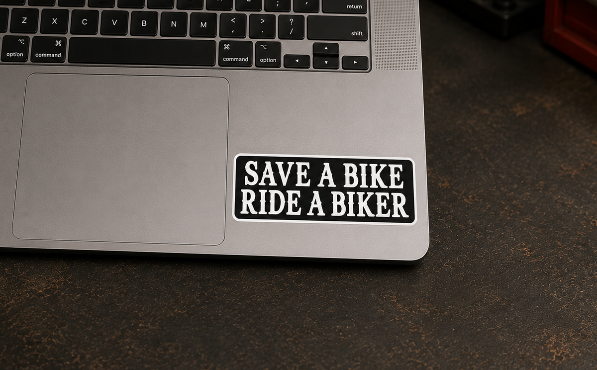 A rectangular black sticker with white text reading 'SAVE A BIKE RIDE A BIKER' applied to the lower right palm rest of a silver laptop, resting on a dark textured tabletop.