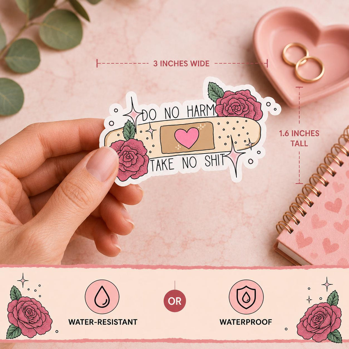 A hand holds a die-cut sticker featuring a pink rose, a beige bandage with a pink heart, and the text 'DO NO HARM TAKE NO SHIT'. The background is a soft pink flat-lay with eucalyptus leaves, a pink heart-shaped dish with gold rings, and a pink notebook.