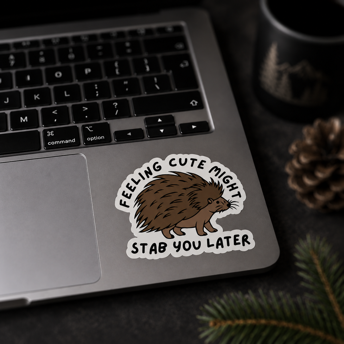 A die-cut vinyl sticker featuring a brown hedgehog illustration and the text 'FEELING CUTE MIGHT STAB YOU LATER' in black, shown applied to the palm rest of a silver laptop. The scene is styled with a dark mug, a pinecone, and evergreen branches on a dark tabletop.