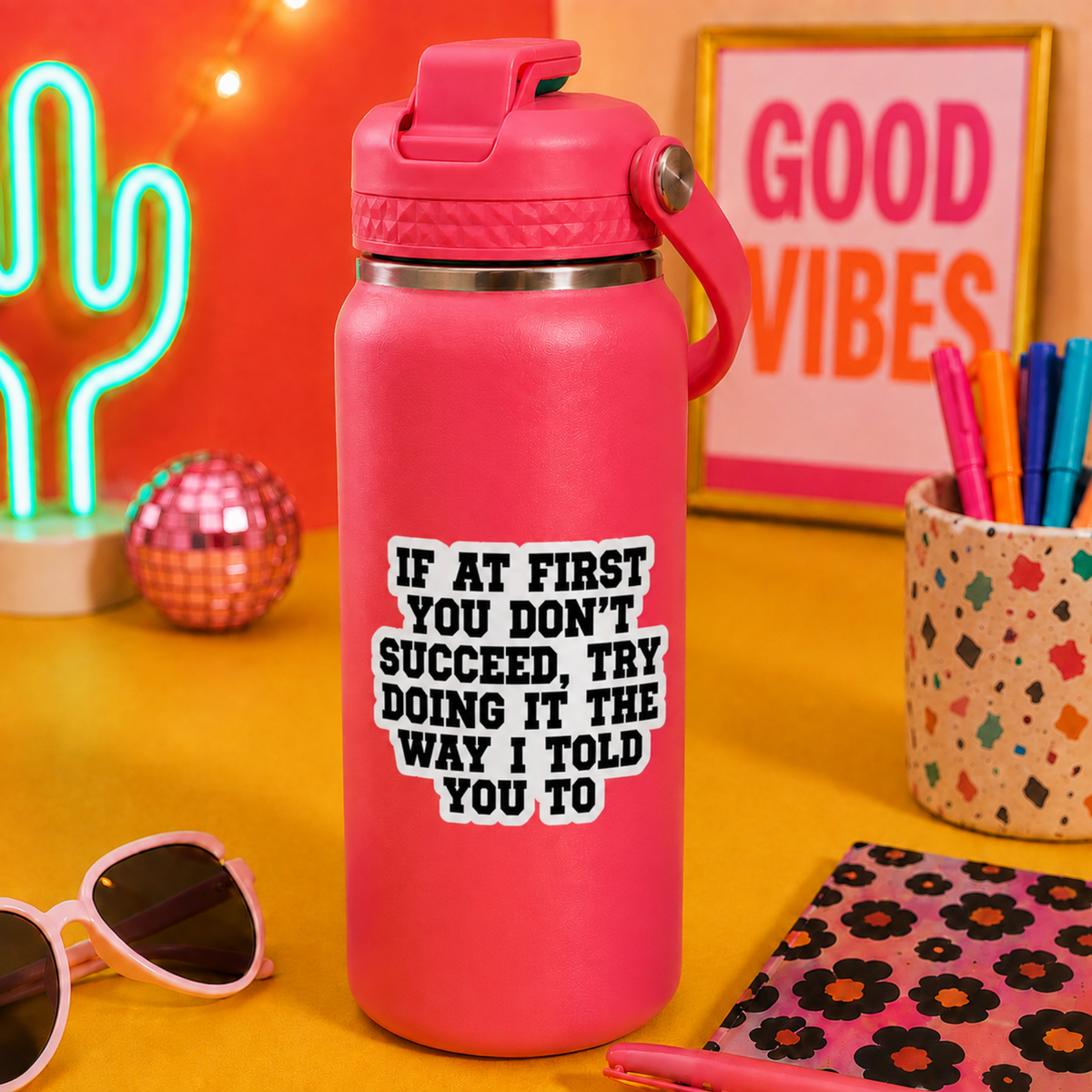 A hot pink insulated water bottle with a matching pink lid and carry loop, standing on a yellow tabletop. The bottle has a bold white text graphic that reads, 'IF AT FIRST YOU DON'T SUCCEED, TRY DOING IT THE WAY I TOLD YOU TO.